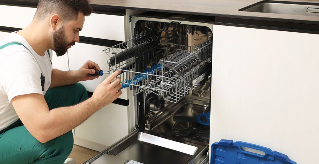 Mains d'un installateur mesurant la largeur d'une niche d'encastrement vide de 60 cm entre deux caissons de cuisine blancs avec mètre ruban métallique, raccords visibles au fond